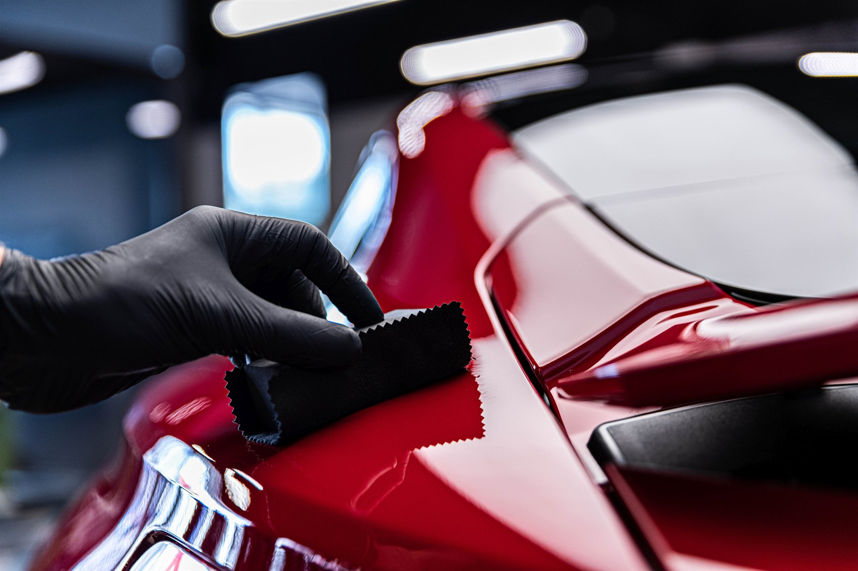 A gloved Detail By Design technician applying ceramic coating to a deep-red sports car in the studio.